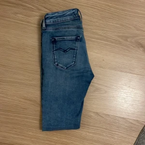 Blå skinny jeans från Replay, W25 L28 - Snygga blå skinny jeans från Replay med klassisk femficksdesign och coola detaljer på bakfickorna. Jeansen har en smal passform och är tillverkade i stretchigt denim för en bekväm känsla. Perfekta till sneakers eller boots.