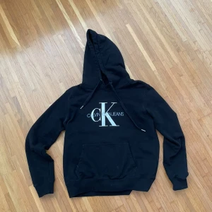 Svart Calvin Klein hoodie med logga - Svart hoodie från Calvin Klein Jeans med stor vit CK-logga på bröstet. Klassisk känguruficka framtill, justerbar huva med snörning och ribbade muddar. Perfekt för en chill och stilren look.