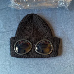 Svart ribbad gogglesmössa - Cool svart stickad mössa med ribbad struktur och inbyggda goggles framtill. Perfekt för dig som vill sticka ut med streetwear-vibe. Mössan är tillverkad i mjukt material och har två stora, runda linser i plast på uppviket.