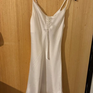Vit långklänning i satin från Bikbok - Supersnygg vit långklänning från Bikbok i glansigt satinmaterial. Klänningen har smala axelband, v-ringning och en snygg snörningsdetalj framtill. Perfekt för dig som vill ha en stilren och elegant look.