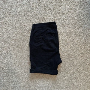 Mörkblå randiga shorts från Selected Homme - Snygga mörkblå shorts från Selected Homme med diskreta vita ränder. Klassisk passform med uppvikta benslut, bälteshällor och fickor både fram och bak. Perfekta för varma dagar när du vill ha en stilren look.