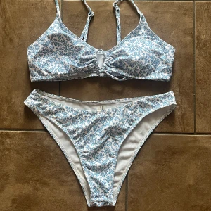 Blommig bikini  i blått och vitt - Superfin bikini från SHEIN i vitt med blått blommönster. Toppen har smala axelband, knytning framtill och en liten cut-out detalj. Bikinitrosan är högt skuren och matchar toppen perfekt. Materialet är mjukt och stretchigt, perfekt för stranden eller poolen. Storlek M, normal i storlek.
