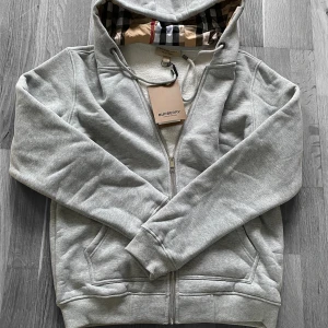 Grå hoodie från Burberry med dragkedja - Snygg ljusgrå hoodie från Burberry med klassiskt rutigt foder i huvan och broderad logga på bröstet. Tröjan har dragkedja, snörning i huvan och mjukt fleecefoder. Perfekt för dig som gillar stilren design med lyxig känsla.