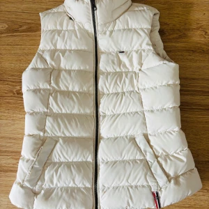 Vit dunväst från Hilfiger Denim - Snygg vit dunväst från Hilfiger Denim med quiltad design och dragkedja framtill. Västen har två fickor med detaljer i rött, vitt och blått samt liten logga på bröstet. 