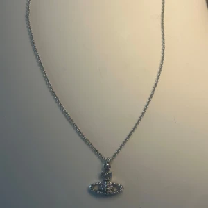 Vivienne Westwood halsband silver/lila - Ikoniskt halsband från Vivienne Westwood i silverfärgad metall med det klassiska orb-hänget dekorerat med ljuslila stenar. Kedjan är tunn och smidig, och hänget har coola nitar och detaljer. Perfekt statement-piece för dig som gillar edgy och trendiga accessoarer.