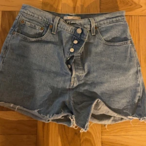 Levi's Ribcage jeansshorts ljusblå - Ljusblå Levi's Ribcage jeansshorts med hög midja och råa, fransiga benslut. Klassisk femficksmodell med knappar framtill och tydliga sömmar. Perfekta för sommaren och ger en avslappnad vibe.