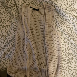 Beige stickad väst med knyt - Säljer en beige stickad väst från kapphal med öppen, virkad design och knytband framtill. Västen har en luftig känsla och fina virkade detaljer längs kanterna. Perfekt att slänga över en t-shirt eller klänning för en chill look.