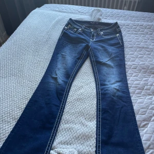 Blå jeans med broderade detaljer från Miss Me - Snygga blå jeans från Miss Me med broderade detaljer och slitningar på bakfickorna. Byxorna har en bootcut-stil och är dekorerade med vita sömmar som ger en cool kontrast. Perfekta för dig som vill ha en unik look! Inköpta i San Francisco.