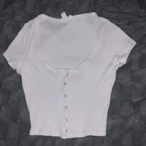 Vit croppad ribbad topp med knappar - Supersöt vit croppad topp med korta ärmar och snygg ribbad struktur. Djup rundad urringning och knappar framtill ger en chill och trendig vibe. Perfekt att matcha med jeans eller kjol för en enkel men stilren look. Och vi kan prata om priset om det behövs! 