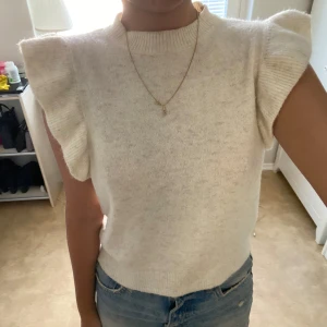 Vit stickad ärmlös tröja med volang - Superfin vit stickad tröja med volangdetaljer på axlarna. Tröjan är ärmlös och har en ribbad halsringning. Perfekt att styla med jeans eller kjol för en fräsch look. Mjuk och skön, passar både till vår och sommar.