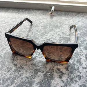 Bruna fyrkantiga solglasögon Vasuma - Snygga solglasögon från Vasuma med markerad fyrkantig form och bruntonade glas. Bågarna är i en cool mix av svart och brun sköldpaddsmönster. Designade i Sverige och tillverkade i Italien. Perfekta för dig som vill sticka ut med en unik look.