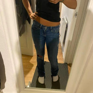 Blå bootcut jeans Hollister W25 L32 - Snygga blå bootcut jeans från Hollister med låg midja och slitningar på ena benet. Jeansen har klassiska bakfickor med broderade detaljer och är gjorda i stretchigt denim för skön passform. Perfekta för dig som gillar en avslappnad och trendig look.