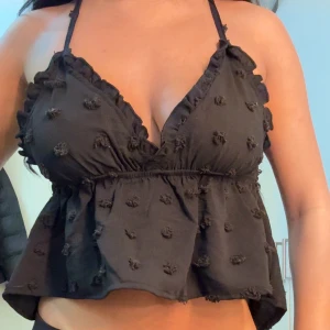 Svart linnetopp med volang och broderi - Supersöt svart linnetopp med tunna korsade axelband och volangkant. Toppen har små broderade detaljer över hela tyget och en lätt v-ringning. 