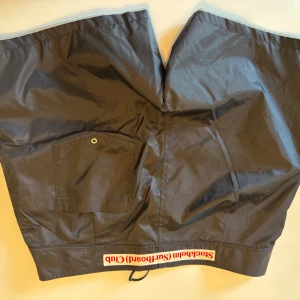 Svarta shorts Stockholm Surfboard Club - Svarta shorts från Stockholm (Surfboard) Club med meshfoder och bakficka med metallöljet. Snygg detalj med rödvit logotyp längs benet och dragsko i nederkant. Perfekta för strand eller chill dagar.