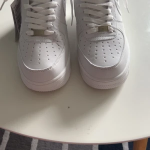 Nike Air Force 1 helvita sneakers - Klassiska Nike Air Force 1 sneakers i helvitt skinn med perforerad tå, vit sula och vita skosnören. Snygg och tidlös modell med låg profil och rund tå, perfekt för dig som vill ha en clean och fräsch look. Ikonisk Nike-logga på sulan och metallbricka vid snörningen.                                           Priset är diskuterbart!