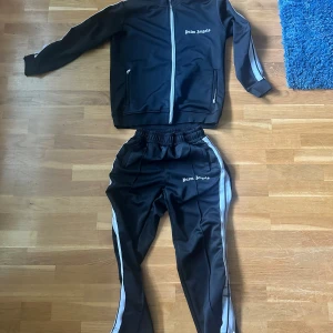 Palm Angels svart träningsset - Snyggt svart träningsset från Palm Angels med vita ränder längs ärmar och ben. Jackan har dragkedja och två fickor med zip, byxorna har resår i midjan och logga på både jacka och byxa. Storlek: Medium