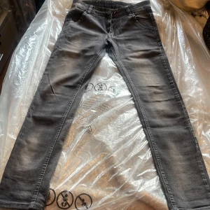 Grå jeans från Sibao Long, stl 31 - Snygga grå jeans från Sibao Long i storlek 31. Jeansen har en tvättad look med ljusare partier och klassisk femficksdesign. Detaljer som färgade sömmar och en liten logga på bakfickan ger extra stil. Perfekta för dig som gillar en avslappnad men trendig vibe.