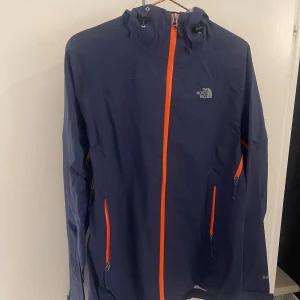 Blå vindjacka från The North Face - Snygg marinblå vindjacka från The North Face med orange dragkedjor och huva. Jackan är tillverkad i slitstark Gore-Tex och har flera praktiska fickor med dragkedja. Perfekt för dig som gillar att vara ute oavsett väder.