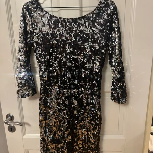 Svart paljettklänning med öppen rygg - Glittrig svart kortklänning täckt av paljetter i silver och guld. Klänningen har trekvartsärm och en snygg öppen rygg med dragkedja bak. Perfekt för dig som vill sticka ut och glänsa på festen!