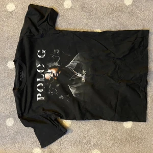 Svart Polo G t-shirt med tryck - Svart t-shirt från Polo G med stort tryck av artisten på framsidan. Klassisk rund hals och korta ärmar. Skön passform och tillverkad i mjuk bomull, perfekt för dig som gillar streetwear och vill sticka ut med en snygg grafisk tee.