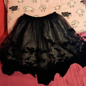 Svart halloween kjol med fladdermusmotiv (barn) - Cool svart kortkjol i tyll med oregelbunden kant och broderade fladdermöss längs fållen. Kjolen har en voluminös look och resår i midjan för skön passform. Perfekt för dig som gillar alternativ stil eller vill sticka ut. Passar även perfekt för halloween!!