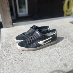 Svarta och vita sneakers från Diesel - Snygga svarta sneakers från Diesel med vita detaljer och logga på plösen. Skorna har en låg profil, snörning och cool röd sula. Tillverkade i skinn och mocka för en schysst mixad look. Perfekta för dig som gillar streetstyle och vill sticka ut lite extra.