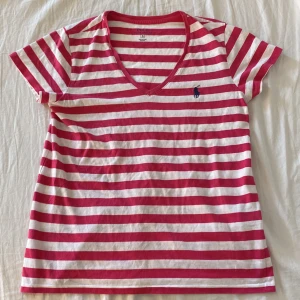 Randig t-shirt från Polo Ralph Lauren - Snygg t-shirt från Polo Ralph Lauren i mörk rosa och vitt med klassiska horisontella ränder. T-shirten har v-ringning, korta ärmar och en broderad marinblå logga på bröstet. Tillverkad i mjuk bomull, perfekt för en avslappnad och stilren look.tröjan ser röd ut på bilden men den är mörkrosa i verkligheten 💖😍
