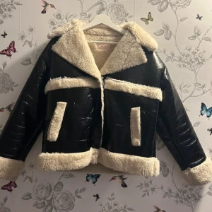 Svart teddyjacka från Nelly, stl 38 - Säljer en svart teddyjacka från Nelly i storlek 38. Jackan har glansig svart utsida och mjukt, beige teddyfoder på krage, ärmslut, fickor och nederkant. Stängs med dragkedja framtill och har en oversized look. Perfekt för kalla dagar när du vill vara både varm och snygg.