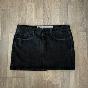 Svart jeanskjol från Disciplin - Snygg svart jeanskjol från Disciplin med klassisk femficksdesign, bälteshällor och coola kontrastsömmar. Kjolen är kort och har en rak passform som ger en avslappnad vibe. Perfekt att matcha med sneakers eller boots för en trendig look.