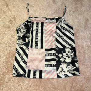 Victoria's Secret linnetopp öppen rygg - Snygg linnetopp från Victoria's Secret med tunna axelband. Öppen rygg. Toppen har ett patchworkmönster med svarta och vita ränder, rosa detaljer och blommor. Materialet är glansigt och tunt, perfekt för att sticka ut. Loggan syns tydligt i designen. Skulle tro att den är xxs eller xs, kort i längd