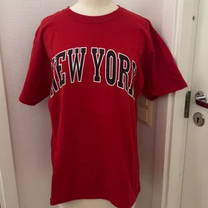 Röd New York t-shirt Fruit of the Loom - Snygg röd t-shirt från Fruit of the Loom med stort svartvitt 'NEW YORK'-tryck över bröstet. Köpt i USA. Ny men tvättad. Medium herr men funkar på tjejer med. Mjuk i bomull. Perfekt för en avslappnad streetstyle-look. Hämtas i Hägersten eller skickas.  