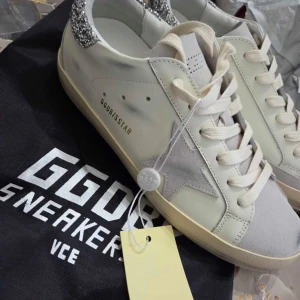 Vita GGDB sneakers med glitterdetalj - Snygga vita sneakers från GGDB med grå mockadetaljer och stjärna på sidan. Hälkappan är täckt av silverglitter för extra edge. Skorna har snörning och en klassisk platt sula. Perfekta för dig som gillar en trendig och unik look.