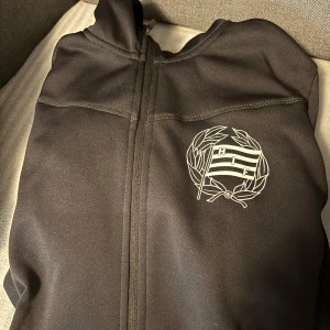 Svart sportig jacka med HIF-logga - Svart sportig jacka med dragkedja och HIF-logga på bröstet. Jackan har huva och är tillverkad i ett mjukt syntetmaterial som passar perfekt för träning eller chill. Enkel och stilren design med diskreta sömmar och hel dragkedja framtill.