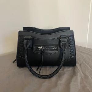 Crossbody bag ginatricot - Snygg svart handväska i skinnimitation med två rejäla handtag och silverfärgade metalldetaljer. Väskan har en dragkedja framtill som är ett fack och ett rymligt huvudfack med innerficka, varav en har dragkedja. Sidorna har dekorativa sömmar och väskan har en stilren, boxig form. Tillkommer ett tillhörande långt band. Väskan är använd väldigt få gånger! Den är slutsåld på hemsidan 