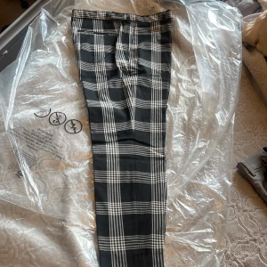 Svarta rutiga kostymbyxor herr 33 - Snygga svarta kostymbyxor med vitt rutmönster. Byxorna har klassisk passform, bälteshällor och knappstängning. Materialet är en mix av polyester, viskos och lite elastan för extra komfort. Perfekta för dig som gillar stilrena och trendiga byxor.
