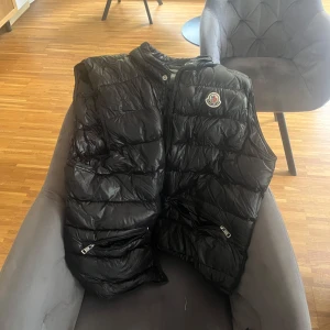 Svart dunväst från Moncler - Snygg svart dunväst från Moncler med glansig finish och klassisk logga på bröstet. Västen har ståkrage, två fickor med dragkedja och quiltad design. Perfekt att slänga över en hoodie eller tröja när det är kyligt ute.