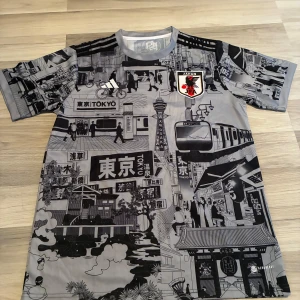 Japan Tokyo Adidas fotbollströja L - Unik fotbollströja från Adidas med Japan-tema och Tokyo-motiv i svart och grått. Tröjan har korta ärmar, coola grafiska tryck med stadsmiljö, tåg och japanska tecken. Märkt med Japan Football Association-emblem och tillverkad i lätt polyester för maximal komfort.