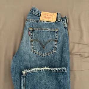 Levi's 501 blå jeans W34 L32 - Klassiska Levi's 501 jeans i blå denim med raka ben och knappgylf. Snygg vintagekänsla med tydliga sömmar och ikonisk läderpatch bak i midjan. Perfekta för dig som gillar tidlös stil och vill ha ett par jeans som funkar till allt. 