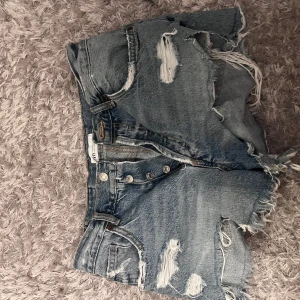 Blå slitna jeansshorts från Zara - Säljer ett par blå jeansshorts från Zara med snygga slitningar och fransig kant. Shortsen har hög midja, knappar framtill och klassisk femficksdesign. Perfekta för en chill sommarlook.