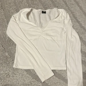 Vit långärmad croppad topp - Snygg vit långärmad topp från Bik Bok med croppad passform och v-ringning. Toppen har en lätt rynkad detalj framtill som ger en cool touch. Perfekt att matcha med jeans eller kjol för en trendig look.