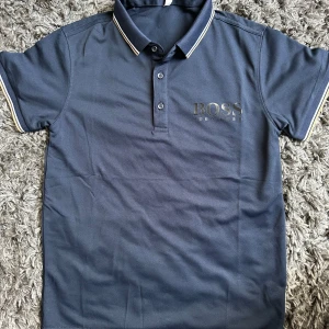 Blå pikétröja från Hugo Boss - Snygg marinblå pikétröja från Hugo Boss med klassisk krage och knäppning framtill. Tröjan har vita och beige ränder på krage och ärmslut samt stor BOSS-logga på bröstet. Perfekt för en stilren och sportig look.