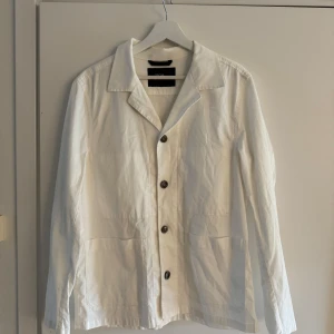 Vit overshirt från Oscar Jacobson - Stilren vit overshirt från Oscar Jacobson med klassisk krage, knäppning framtill och två stora fickor. Tillverkad i bomull för en skön och lätt känsla. Perfekt att slänga på sig över en t-shirt för en clean look.