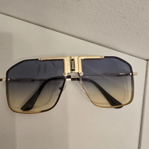 Guldiga fyrkantiga solglasögon - Stilrena solglasögon med fyrkantiga linser och guldig metallbåge. Glasen har en snygg toning från mörkgrått till ljust beige, vilket ger en modern och cool vibe. Svarta skalmar och detaljerad brygga framtill. Perfekta för dig som vill sticka ut.