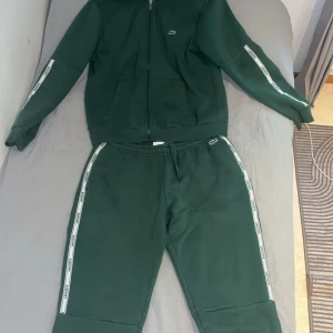 Lacoste dress med dragkedja - Mörkgrön dress från Lacoste med vit logotypband längs ärmarna och klassisk krokodil på bröstet. Tröjan har huva med snörning, dragkedja framtill och två fickor. Perfekt för en sportig och chill stil. Tvättas innnan ja skickar den