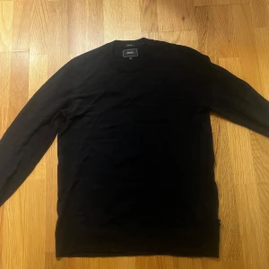 Svart merinotröja från Boomerang - Snygg svart tröja från Boomerang i 100% merinoull. Klassisk rund halsringning och cool detalj med knappar på ena axeln. Långärmad och sitter riktigt bra, perfekt till jeans eller chinos. Enkel och stilren design som funkar året runt. Pga att det är merinoull går det lika bra att använda plagget som snygg tröja till vardagen eller på skidsemestern under skaljackan.