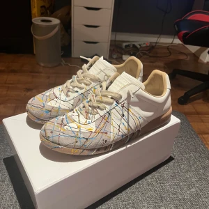 Maison margiella Gats - Maison Margiela sneakers i vitt skinn med unika färgstänk i blått, gult, rött och svart. Klassisk låg modell med beige sula och snörning. Snygg och konstnärlig vibe, perfekt för dig som vill sticka ut med dina skor. 