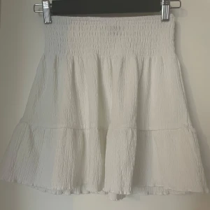 Vit volangkjol från Gina Tricot - Supersöt vit kortkjol från Gina Tricot i storlek XXS. Kjolen har smockad midja och volanger som ger en flowy och luftig känsla. Perfekt för varma dagar och enkel att styla med crop tops eller oversized t-shirts.