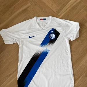 Inter Milan vit bortatröja Nike - Inter Milan bortatröja från Nike i vitt med diagonal pixelmönstrad blå och svart rand över framsidan. Broderat klubbmärke och stjärna på bröstet, blå Nike-logga och V-ringning. Tillverkad i lätt och ventilerande polyester, perfekt för match eller träning.