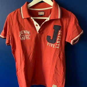 Röd pikétröja från Jack & Jones - Röd piké från Jack & Jones med vita och marinblå detaljer på krage och ärmslut. Stora tryck och broderier framtill med text och siffror. Klassisk krage med knappar och korta ärmar. Perfekt för en sportig och avslappnad stil.