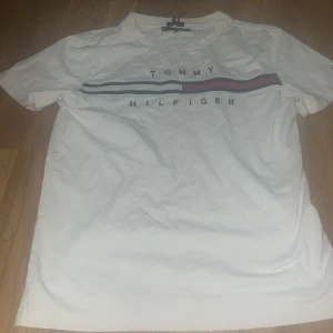 Vit Tommy Hilfiger t-shirt med tryck - Vit t-shirt från Tommy Hilfiger med klassiskt tryck över bröstet och ränder i blått och rött. T-shirten har rund hals, korta ärmar och är gjord i mjuk bomull. Perfekt för en clean och stilren look.